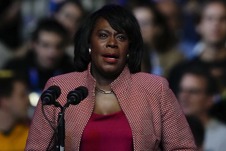 Woman in red shirt and red and white patterned jacket speaks in front of microphone to an audience