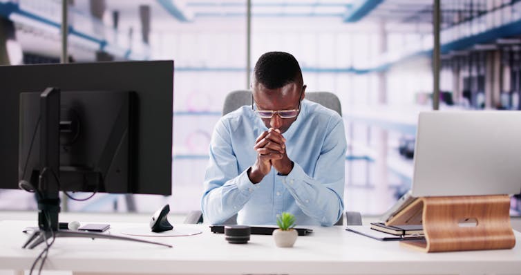 A Black man folding his hands in prayer while seated in front of his desktop computer.