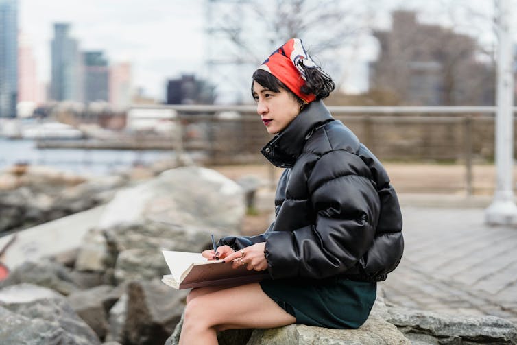 A girl sits on a low wall writing in a notebook.