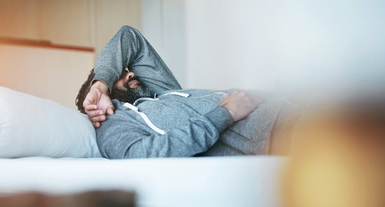 Man in grey sweatshirt lies in bed with arm over head.
