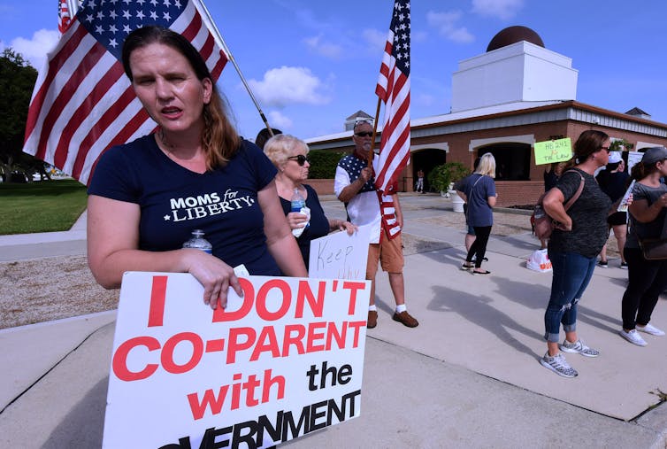 Women stand on a street and hold American flags and a sign that says, 'I don't co-parent with the government.'