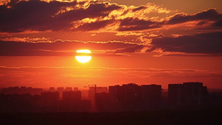 The sunset casts a red glow on the clouds above the evening city skyline with its tall rooftops.