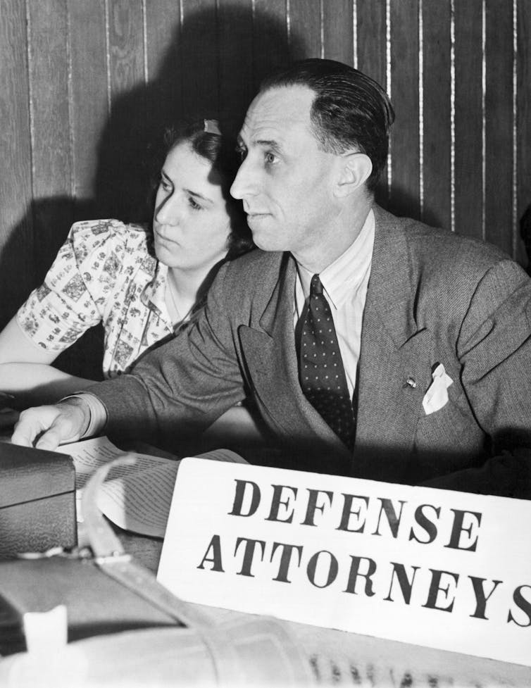 A man with dark hair and in a suit, sitting and listening as a teenage girl leans on his shoulder.