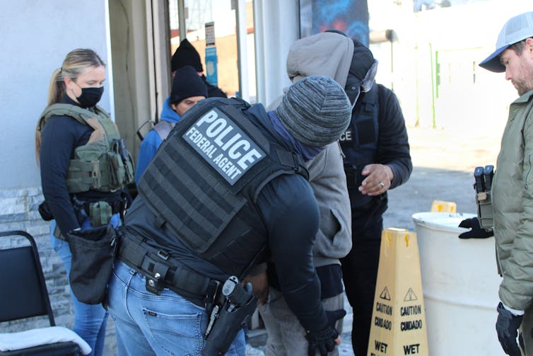 Person with vest labeled 'Police Federal Agent' shown patting down person in jeans and hoodie as other people stand and watch