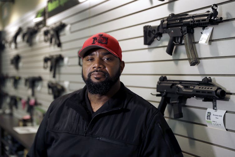A man with a dark beard and red hat posing in front of a wall featuring many guns for sale.