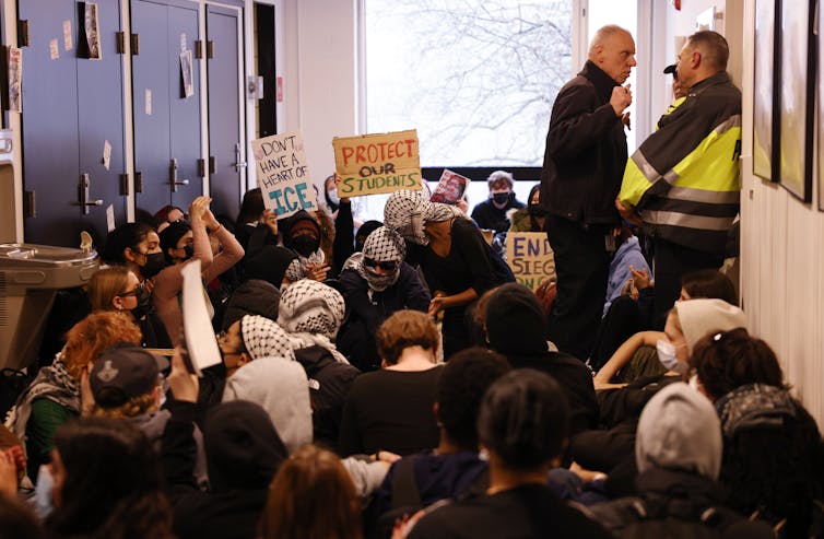 Dozens of students sit in a hallway as police officers stand nearby speaking to each other.