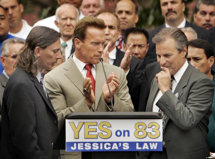 A man appears at a podium with a man to his left and right, and with several people behind the podium.