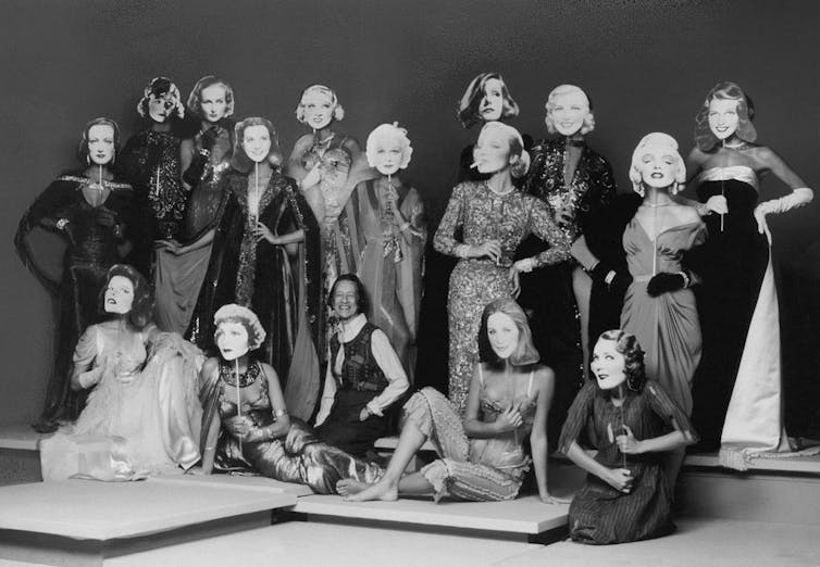 Sixteen woman pose wearing elegant dresses while holding up masks of iconic Hollywood movie stars.