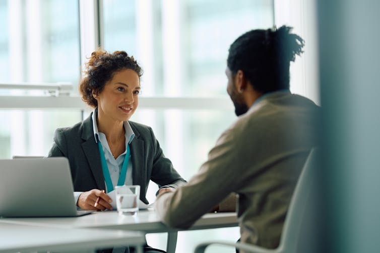 A woman speaks to a man seated across from her at a desk in an office