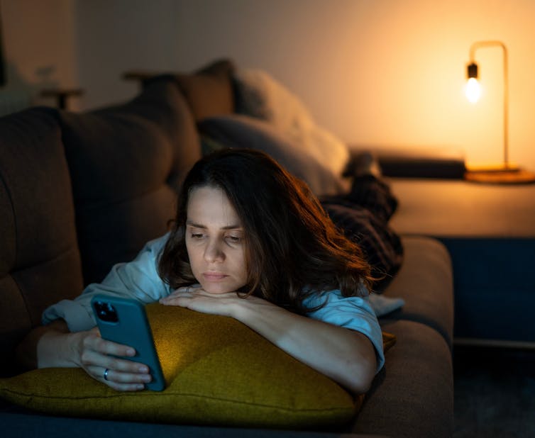 A woman lies on her stomach on a couch, scrolling on her phone and looking distressed, in the evening