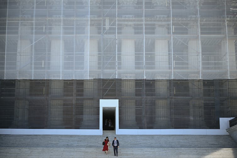 Two people walk down a long set of steps outside of a tall building that is covered in construction facade work.