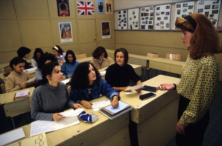 A woman stands and looks at young people seated at a table together with paper in front of them in a dated looking colored photo.