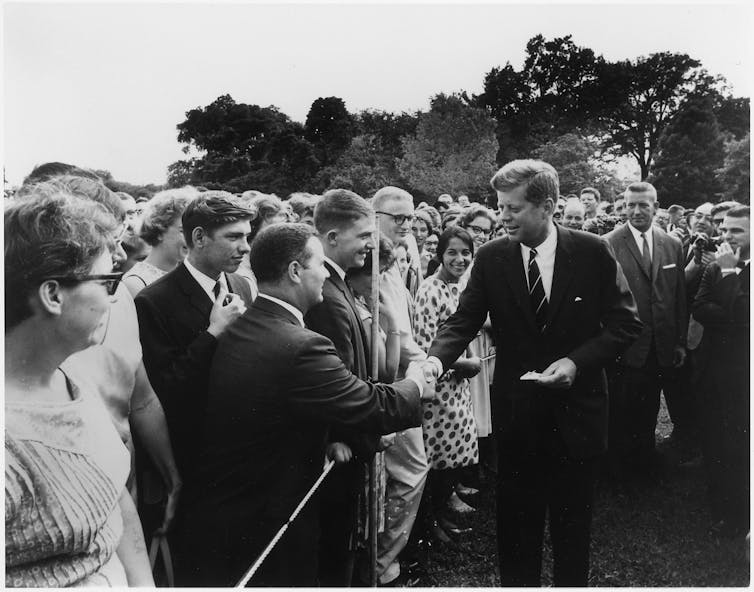 A white man wearing a dark suit shakes hands with a young man in a crowd of people in a black and white photo.