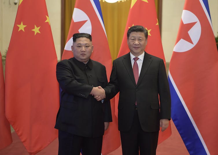 Two men shake hands in front of flags.