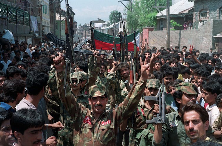 Men in army fatigues hold guns aloft while walking with people on a crowded street.