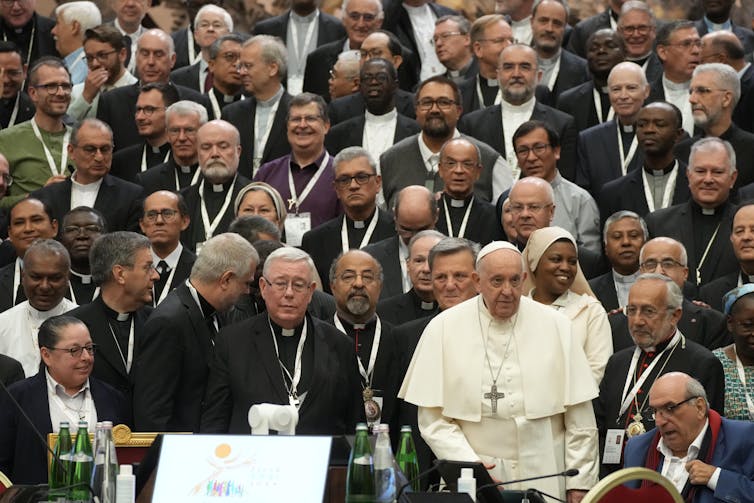 A man in a white priestly robe and a crucifix around his neck stands with several others, dressed mostly in black.