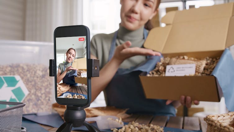 A smartphone on a stand records a woman opening a box and displaying the contents