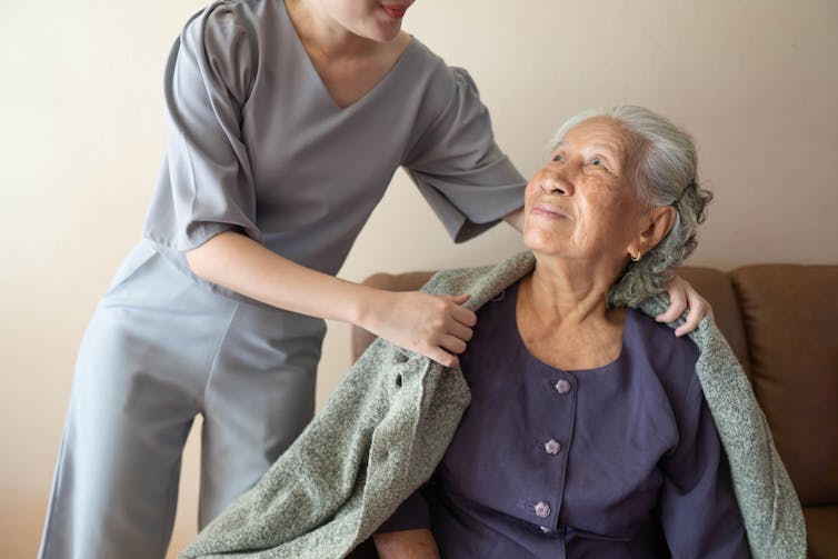 A younger person out of frame putting a sweater over the shoulders of an older woman