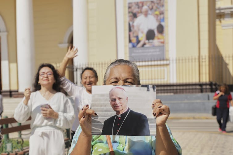 A woman holds a photograph of a man wearing a black clerical coat, while two others stand beside her and wave.