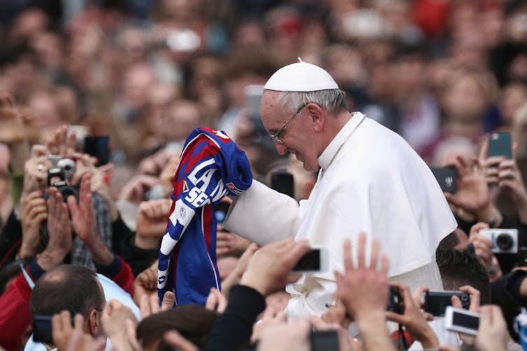 A man in a white cap surrounded by people and clutching a blue and red jersey.