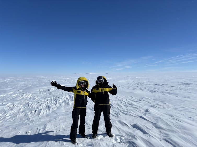 Two people in winter gear stand on a field of white ice with blue sky above them.