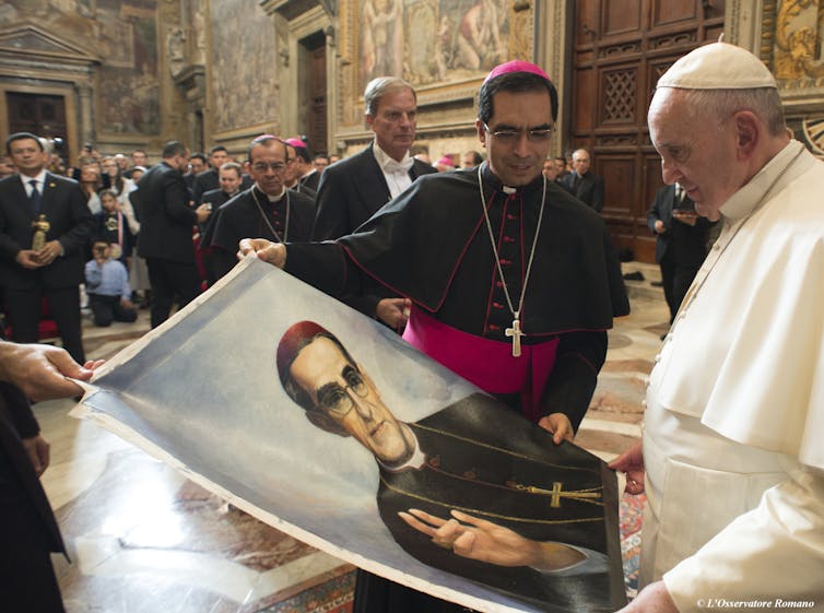 A man in a white robe, cape and skullcap looks at a poster of a priest in a black robe, held by two other men.