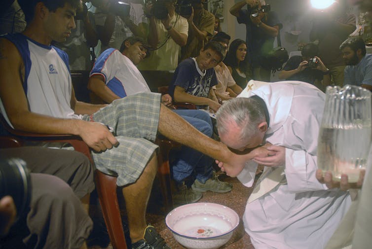 An older man in a white robe kneels on the floor by a small basin as he holds and kisses the foot of a young man in shorts and a T-shirt.