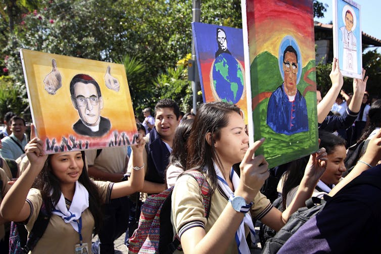 A group of young people walk outside as they hold up brightly colored paintings, each of which depicts a man in priest's robes.