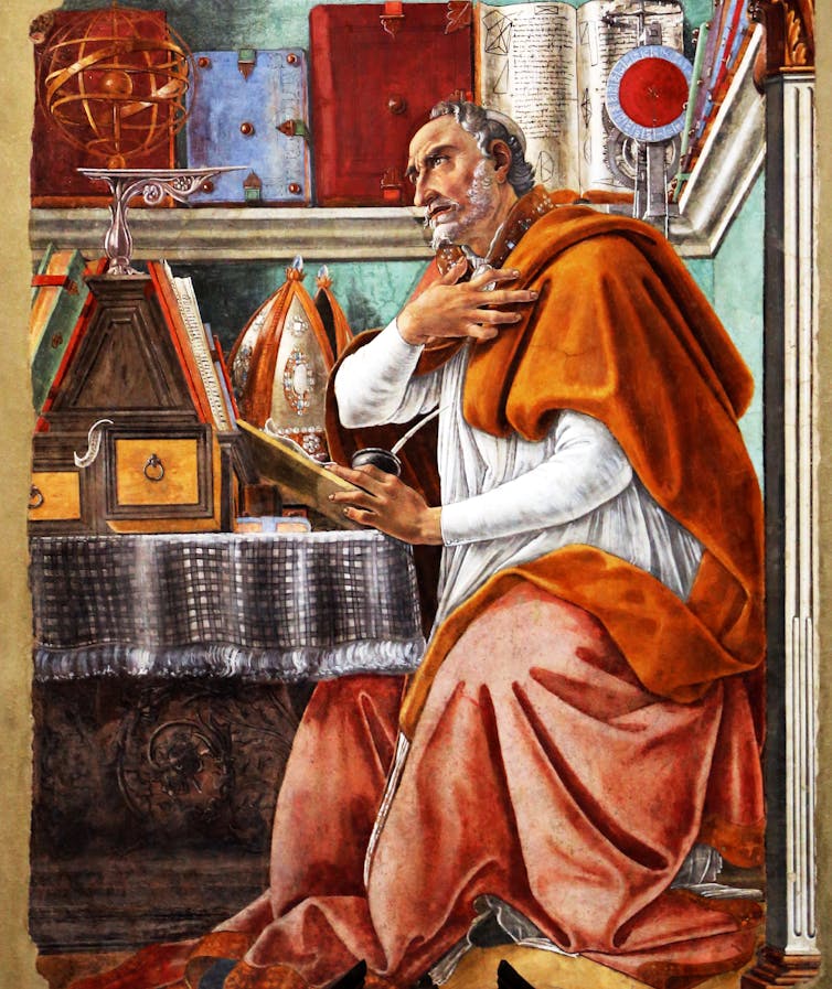 A man in white and saffron robes, seated at a table with an open book in his hand, gazes ahead.