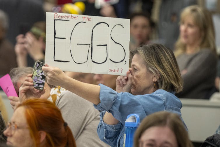 A woman at a large meeting holding a sign that says 'Remember the EGGS, stupid?'