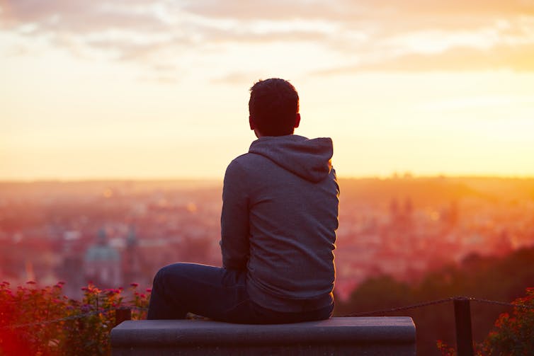 View from behind of a young man sitting alone looking out over the horizon