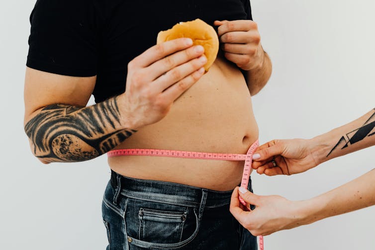 a man with a small belly and tattooed arms eating a burger while hands measure his bare stomach