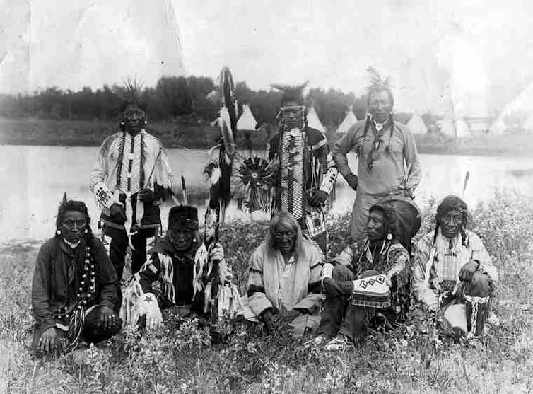 A black-and-white photo of men in Indigenous gala attire. The tall man at back of group is wearing an overall. The white cuffs and all cross-stitch patterns are their own handiwork.